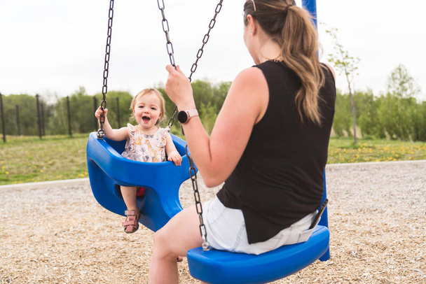 Cute baby girl playing on outdoor playground with mother on swing - Photo, Image