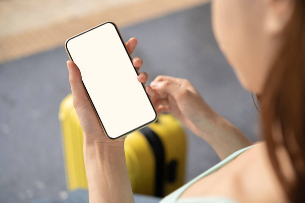 Back view of girl holding smartphone with white blank screen mockup. Traveler with phone at the airport with passport and suitcase. Lifestyle concept with digital technology. - Φωτογραφία, εικόνα