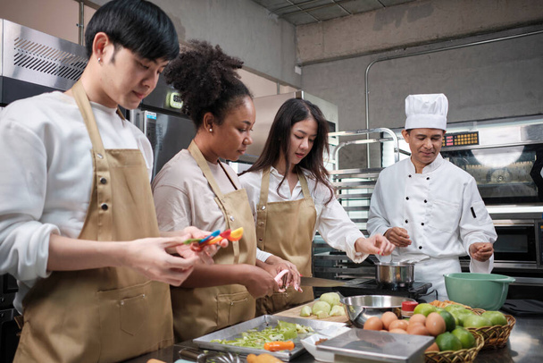 Hobby-Kochkurs, Seniorchef in Kochuniform bringt jungen Kochkursteilnehmern bei, Äpfel zu schälen und zu hacken, Zutaten für Gebäck, Obstkuchen in der Edelstahlküche. - Foto, Bild