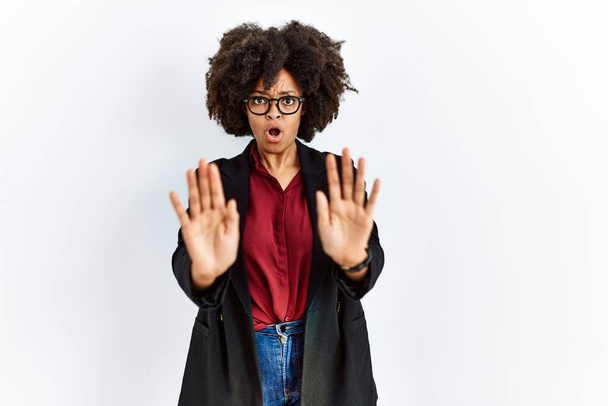 ビジネスジャケットや眼鏡を着用したアフロヘアのアフリカ系アメリカ人女性は、恐怖と嫌な表情で拒否と否定を示す手のひらを離れて移動します。止めて禁止する.  - 写真・画像