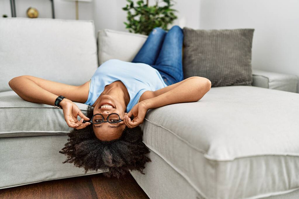 Genç Afro-Amerikan kadın, evinde koltukta kendinden emin bir şekilde yatarken gülümsüyor. - Fotoğraf, Görsel