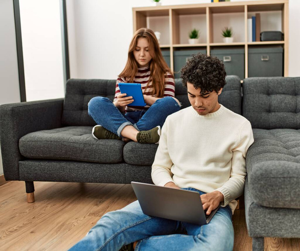 Young couple with serious expression using laptop and touchpad sitting on the sofa at home. - Photo, Image