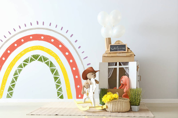Toys, cardboard house and basket with flowers near light wall with painted rainbow - Фото, изображение
