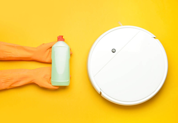 Female hands in rubber gloves, bottle of detergent and robot vacuum cleaner on color background - Photo, Image