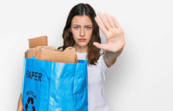 Young beautiful woman holding recycling wastebasket with paper and cardboard with open hand doing stop sign with serious and confident expression, defense gesture  - Фото, изображение