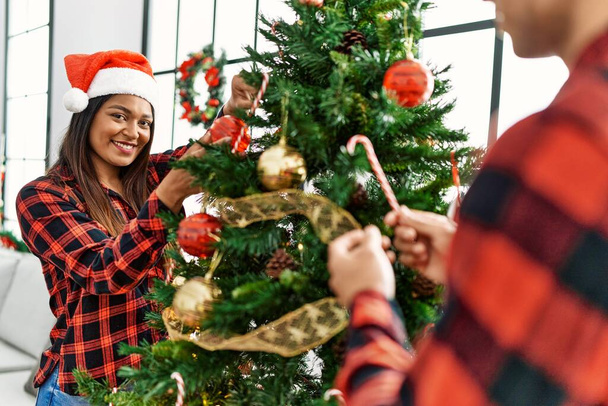 Joven pareja latina sonriendo feliz decorando el árbol de Navidad en casa. - Foto, imagen