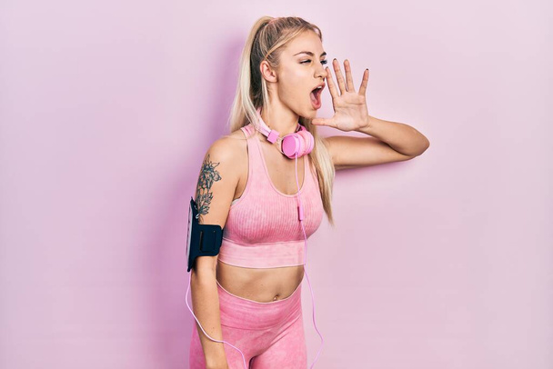 Young beautiful caucasian woman wearing gym clothes and using headphones shouting and screaming loud to side with hand on mouth. communication concept.  - Foto, Imagem