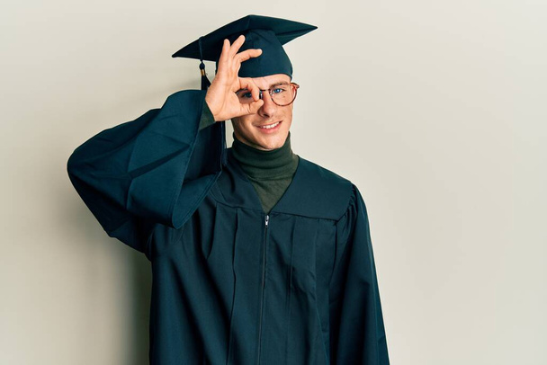 Young caucasian man wearing graduation cap and ceremony robe doing ok gesture with hand smiling, eye looking through fingers with happy face.  - Фото, изображение
