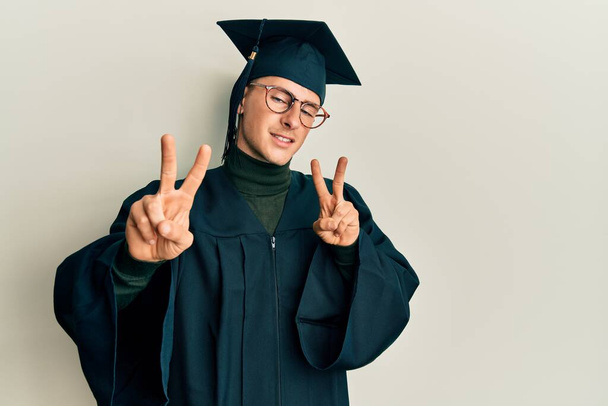 Jeune homme caucasien portant casquette de graduation et robe de cérémonie souriant regardant la caméra montrant les doigts faisant signe de victoire. numéro deux.  - Photo, image