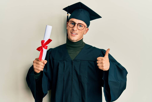 Young caucasian man wearing graduation cap and ceremony robe holding diploma smiling happy and positive, thumb up doing excellent and approval sign  - 写真・画像