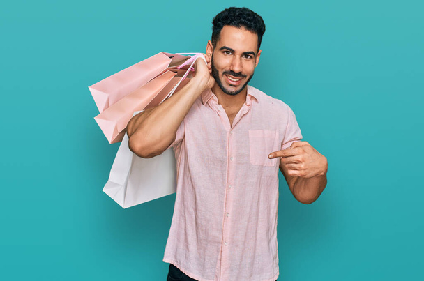 Latynos z brodą trzymający torby na zakupy wskazujące palcem na jednego uśmiechniętego szczęśliwego i dumnego  - Zdjęcie, obraz