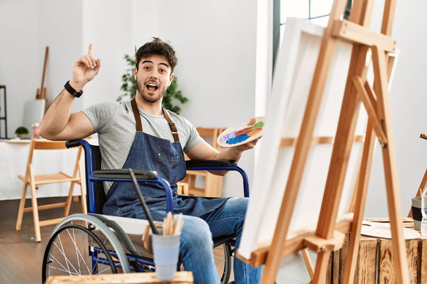 Joven hombre hispano sentado en la pintura en silla de ruedas en el estudio de arte señalando con el dedo hacia arriba con una idea exitosa. exitado y feliz. número uno.  - Foto, imagen