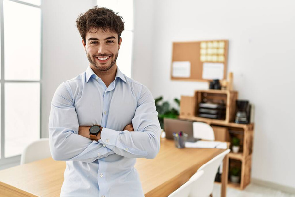 Молодой латиноамериканец-бизнесмен улыбается счастливо стоя со скрещенными руками в офисе. - Фото, изображение