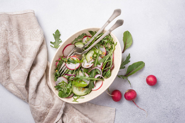 Healthy and tasty vegan food. Top view of light salad with radish, cucumber, onion, fresh green leaves and mustard sause on grey concrete background - Foto, immagini