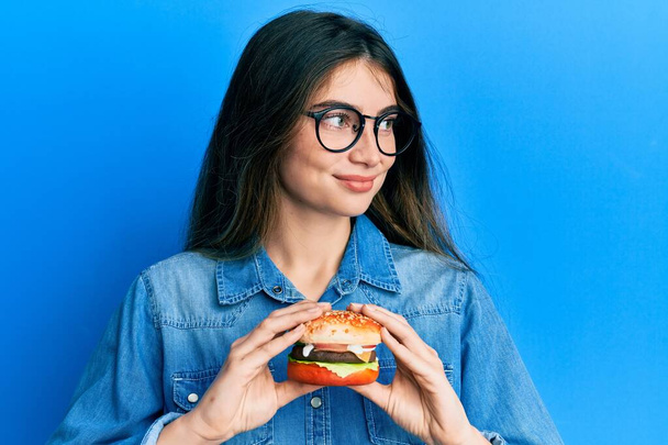 Young caucasian woman eating a tasty classic burger smiling looking to the side and staring away thinking.  - 写真・画像