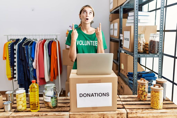 Junge blonde Frau im freiwilligen T-Shirt am Spendenstand steht erstaunt und überrascht auf und zeigt mit erhobenen Armen und Fingern nach oben.  - Foto, Bild