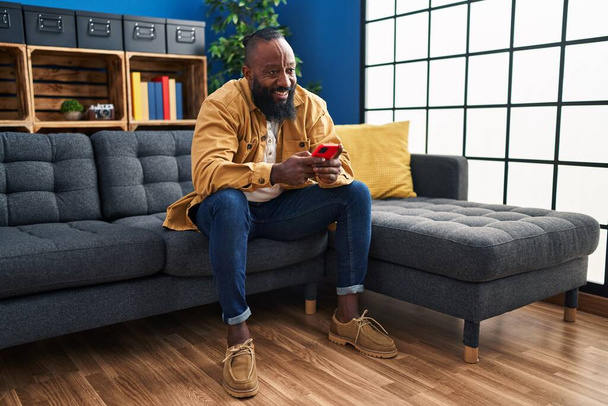Young african american man using smartphone sitting on sofa at home - Fotó, kép