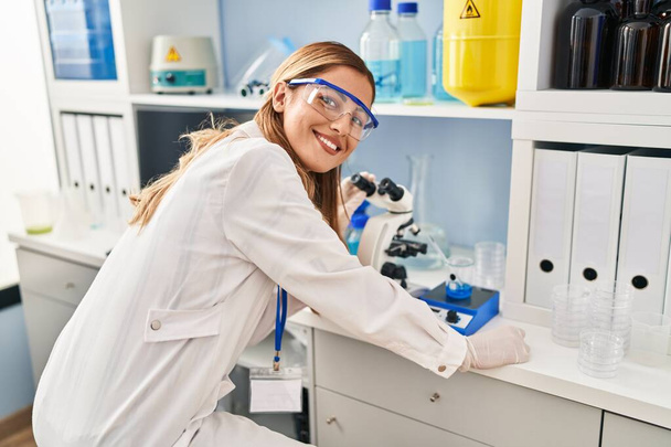 Młoda blondynka w mundurze naukowca używająca mikroskopu w laboratorium - Zdjęcie, obraz