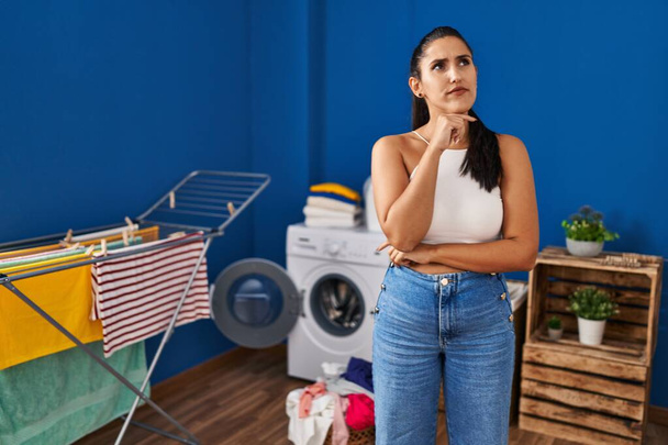 Young hispanic woman at laundry room serious face thinking about question with hand on chin, thoughtful about confusing idea  - Foto, imagen