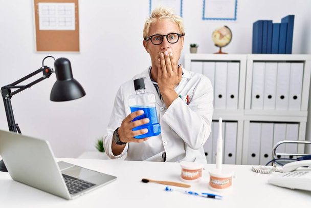 Young blond dentist man holding mouthwash at clinic covering mouth with hand, shocked and afraid for mistake. surprised expression  - Photo, Image
