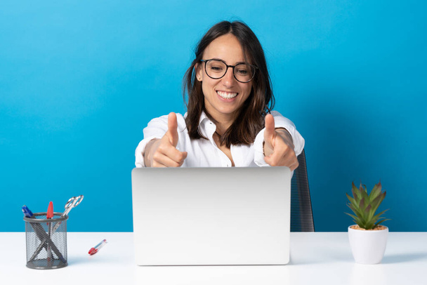Spaanse mooie jonge vrouw maakt ok teken in de voorkant van laptop geïsoleerd op blauwe achtergrond. Kantoormedewerker op videoconferentie - Foto, afbeelding