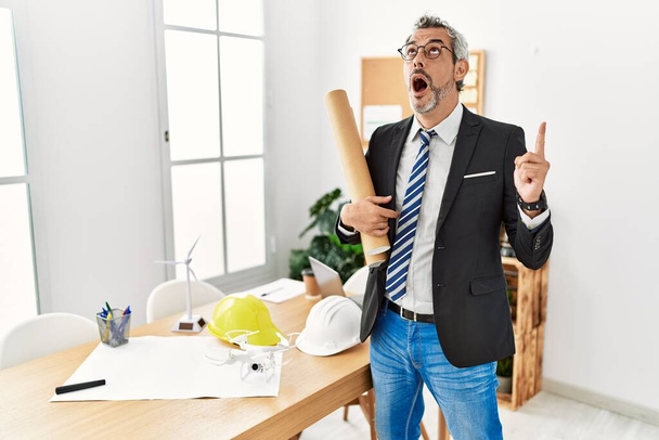 Middle age hispanic business man holding paper blueprints amazed and surprised looking up and pointing with fingers and raised arms.  - 写真・画像