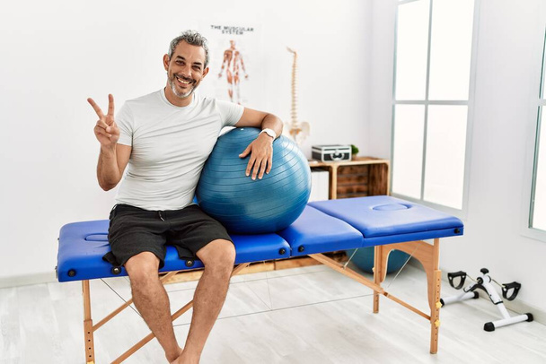 Middle age hispanic man at pain recovery clinic holding pilates ball smiling with happy face winking at the camera doing victory sign with fingers. number two.  - Foto, Imagem