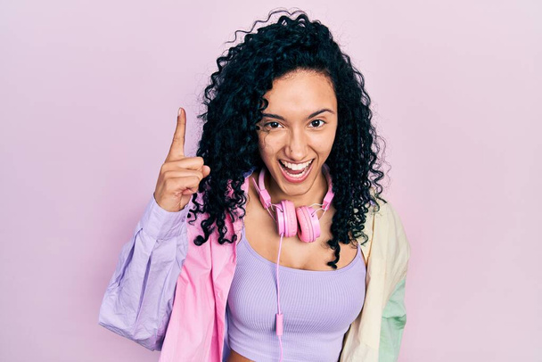 Junge hispanische Frau mit lockigem Haar in Turnbekleidung und Kopfhörern, die mit erhobenem Zeigefinger auf eine erfolgreiche Idee hinweisen. aufgeregt und glücklich. Nummer eins.  - Foto, Bild