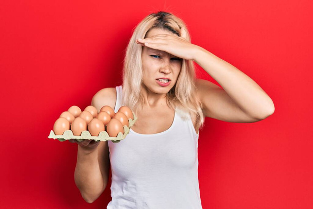 Beautiful caucasian blonde woman holding tray of fresh eggs stressed and frustrated with hand on head, surprised and angry face  - Φωτογραφία, εικόνα