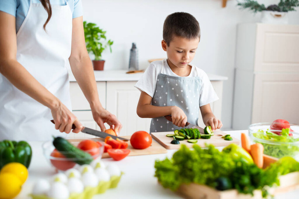 Zoon helpt moeder thuis. Kind kookt met moeder in de witte moderne keuken, het snijden van komkommers voor salade. Samenhorigheidsconcept. Gezond voedsel. - Foto, afbeelding