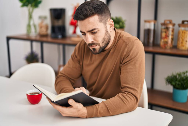 Young hispanic man reading book and drinking coffee sitting on table at home - Photo, Image
