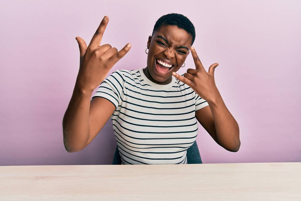 Молодая африканская американка, одетая в повседневную одежду, сидит на столе и кричит с сумасшедшим выражением, делая рок-символ с поднятыми руками. музыкальная звезда. тяжёлая концепция.  - Фото, изображение