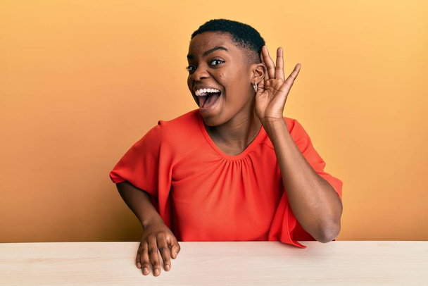 Νεαρή Αφροαμερικανή γυναίκα που φοράει καθημερινά ρούχα κάθεται στο τραπέζι χαμογελώντας με το χέρι πάνω από το αυτί ακούγοντας φήμες ή κουτσομπολιά. έννοια της κώφωσης.  - Φωτογραφία, εικόνα