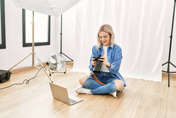 Όμορφη ξανθιά γυναίκα που εργάζεται ως επαγγελματίας φωτογράφος στο στούντιο φωτογραφίας κάθεται στο πάτωμα ελέγχοντας φωτογραφίες στο φορητό υπολογιστή - Φωτογραφία, εικόνα