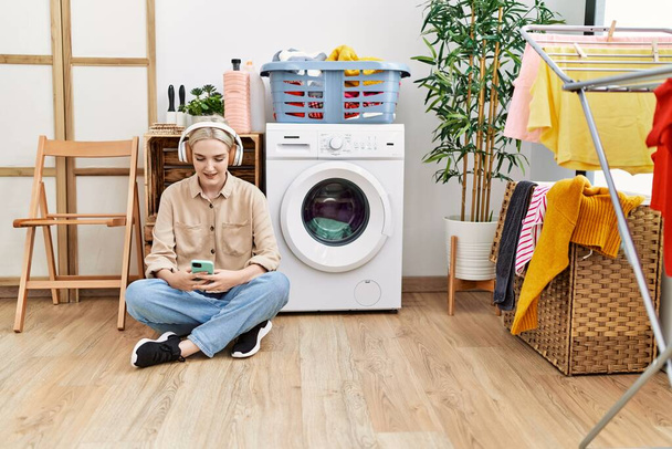 Joven mujer caucásica escuchando música esperando la lavadora en la sala de lavandería - Foto, imagen