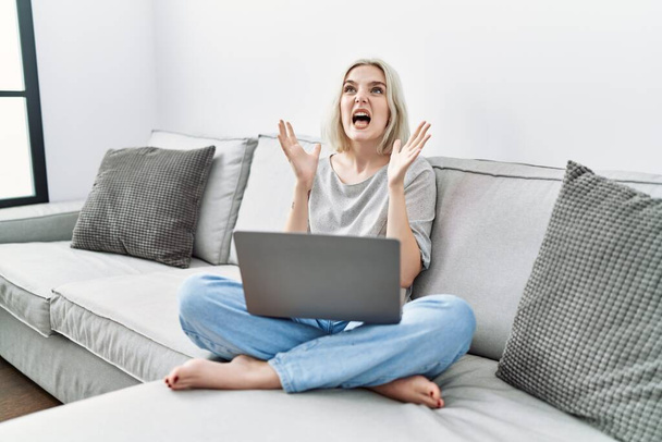 Νεαρή καυκάσια γυναίκα που χρησιμοποιεί φορητό υπολογιστή στο σπίτι κάθεται στον καναπέ τρελή και τρελή φωνάζοντας και φωνάζοντας με επιθετική έκφραση και τα χέρια ψηλά. έννοια απογοήτευσης.  - Φωτογραφία, εικόνα