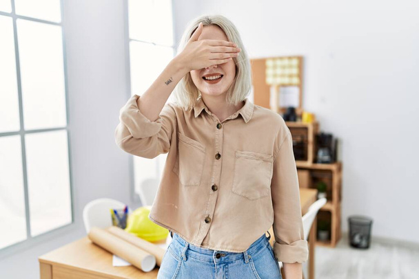 Young beautiful caucasian woman at construction office smiling and laughing with hand on face covering eyes for surprise. blind concept.  - Foto, immagini