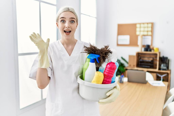 Young caucasian woman wearing cleaner uniform holding cleaning products cleaning office celebrating victory with happy smile and winner expression with raised hands  - Valokuva, kuva