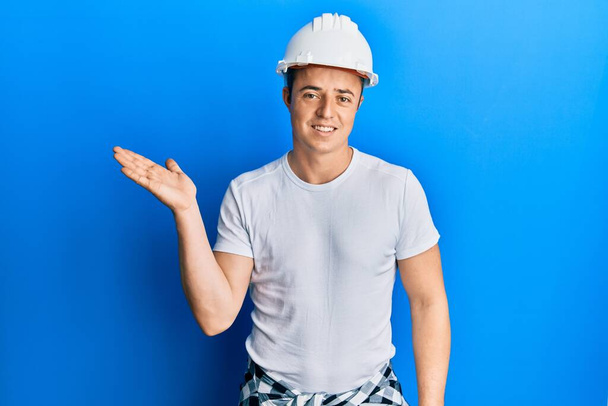 Beau jeune homme portant uniforme de constructeur et casque dur souriant présentation joyeuse et pointant avec la paume de la main en regardant la caméra.  - Photo, image