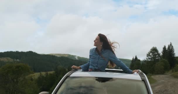 bela menina de cabelos compridos inclinou-se para fora da escotilha de um carro estacionado nas montanhas. Olha em volta da floresta verde e da natureza. Tiro médio, geralmente - Filmagem, Vídeo