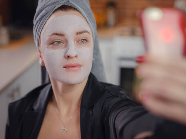 Młoda kobieta w kremowej masce błotnej na twarzy robi sobie selfie telefonem. blogerka piękności pokazująca jej wygląd przez smartfon podczas zabiegów SPA. Spa i Wellness. - Zdjęcie, obraz
