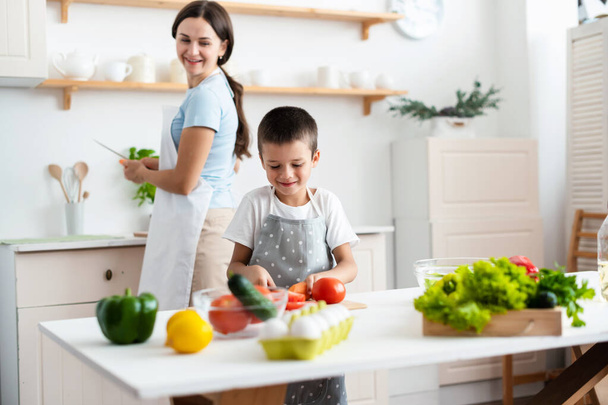 Cheerful mother proud her self-dependent son. Child unaided is cooking salad at kittchen. Healthy food concept. - Foto, imagen