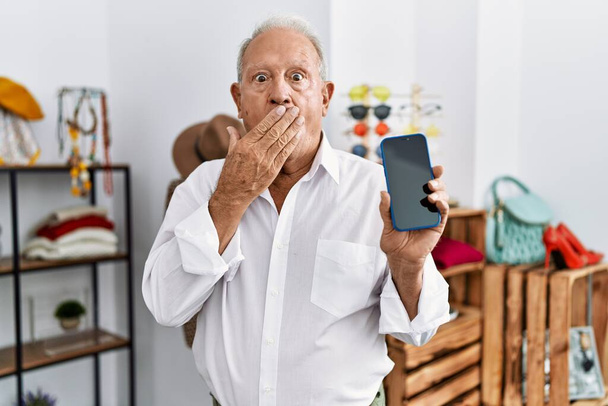 Senior man holding smartphone at retail shop covering mouth with hand, shocked and afraid for mistake. surprised expression  - Photo, Image
