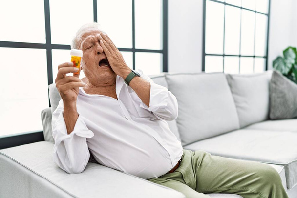 Hombre mayor sosteniendo pastillas bostezando cansado cubriendo media cara, ojo y boca con la mano. cara duele en el dolor.  - Foto, imagen