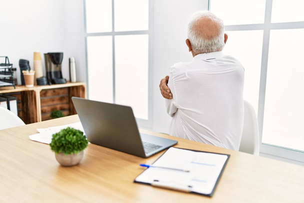 Старший человек, работающий в офисе, используя ноутбук, обнимающий себя счастливым и позитивным с обратной стороны. любовь к себе и забота о себе  - Фото, изображение