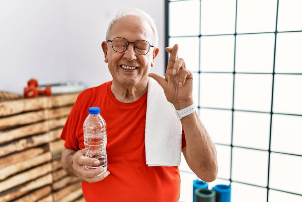 Старший чоловік у спортивному одязі та рушнику у спортзалі жестикулює палець схрещеним усміхненим з надією та закритими очима. удача і забобонна концепція
.  - Фото, зображення