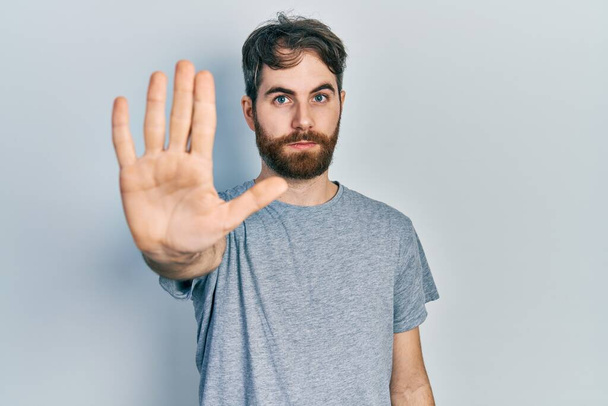 Caucasian man with beard wearing casual grey t shirt doing stop sing with palm of the hand. warning expression with negative and serious gesture on the face.  - Photo, image