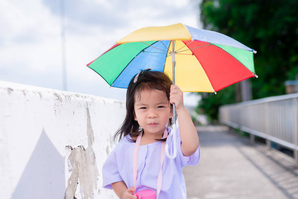 Portrait image of 4-5 years old. Cute Asian girl walks with umbrella in bright sun, to protect her from scorching sunny. In summer time. Kindergarten students are walking home after school. Copy space - Foto, Imagem