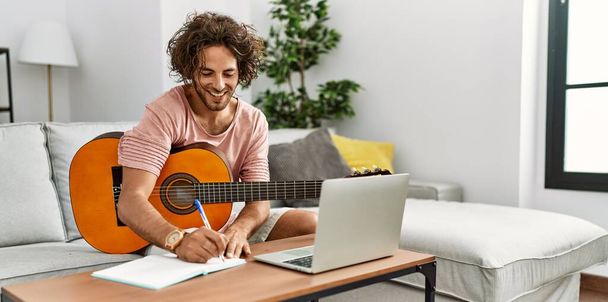 Joven compositor hispano componiendo canción usando guitarra y laptop en casa. - Foto, imagen
