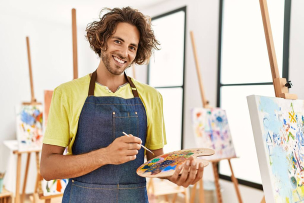 Young hispanic artist man smiling happy holding paintbrush and palette at art studio. - Photo, image